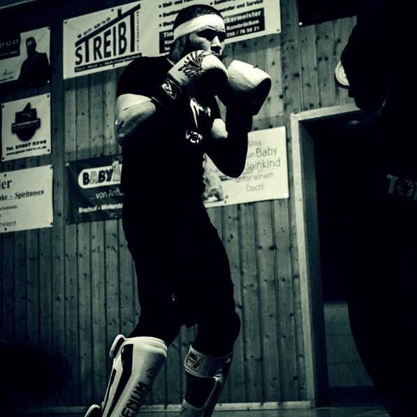 Karol Huber Kickboxen Ein Wettkämpfer trainiert mit Boxhandschuhen und Schienbeinschoner in einem schlichten Trainingsraum.